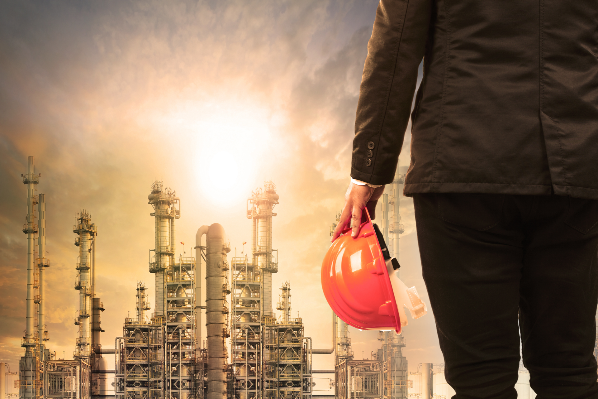 engineering man with safety helmet standing in industry estate against sun rising above oil refinery plant